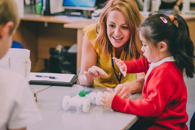Teacher is sitting at the table in her classroom with her primary school students. They have built a car from recycled objects and crafts equipment and are testing that it works.